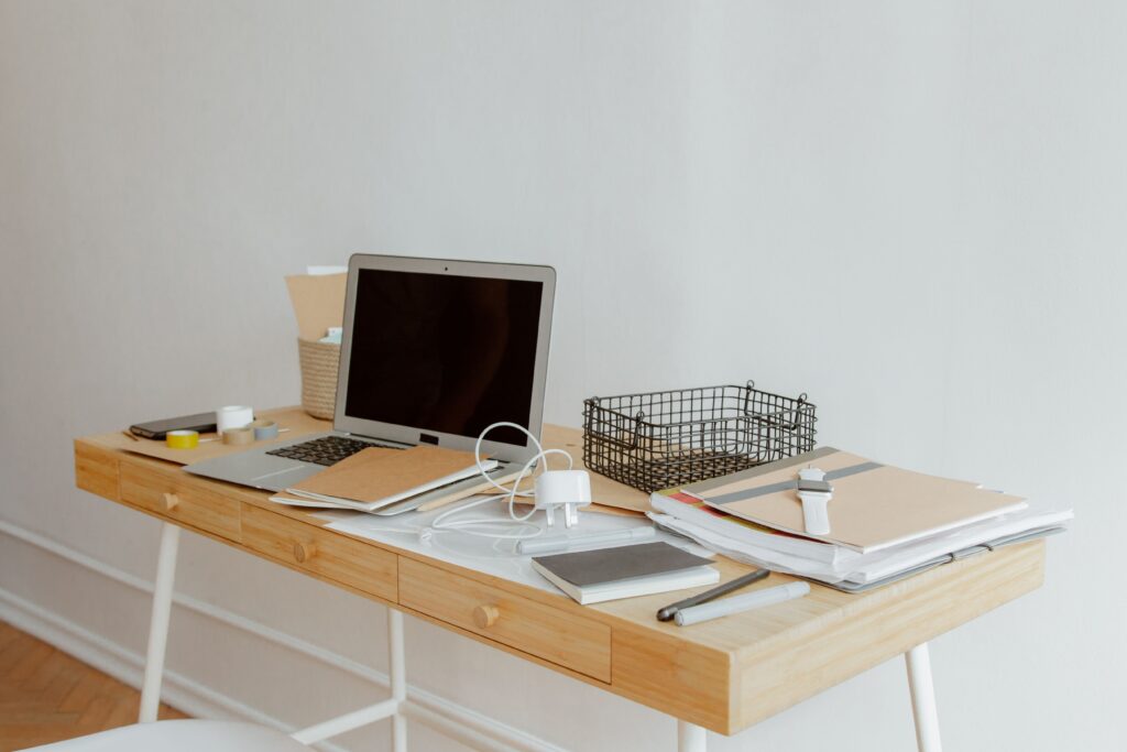 Desk | gearandcode Desk cluttered with an open laptop white cables and notebooks on a light wooden desk against a pale wall