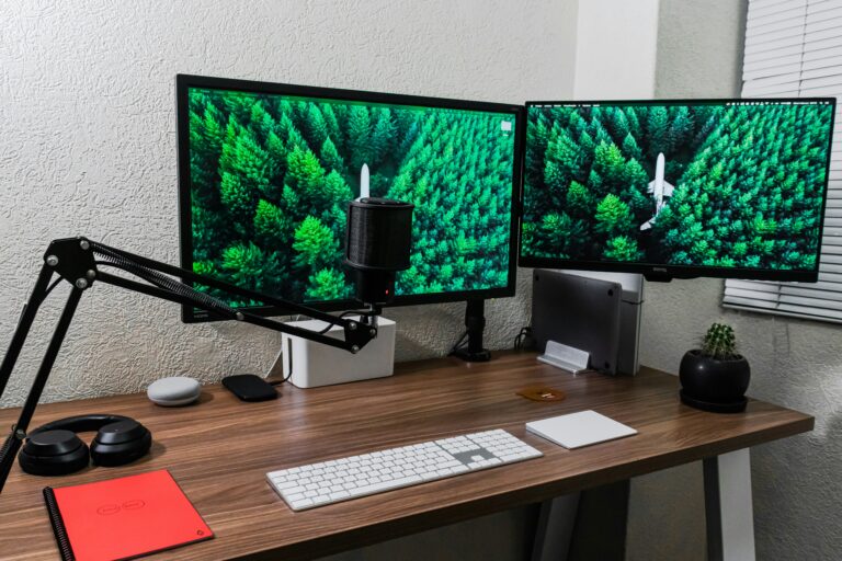Dual monitor computer setup on a wooden desk with a microphone keyboard and a bright red notebook small cactus on the right