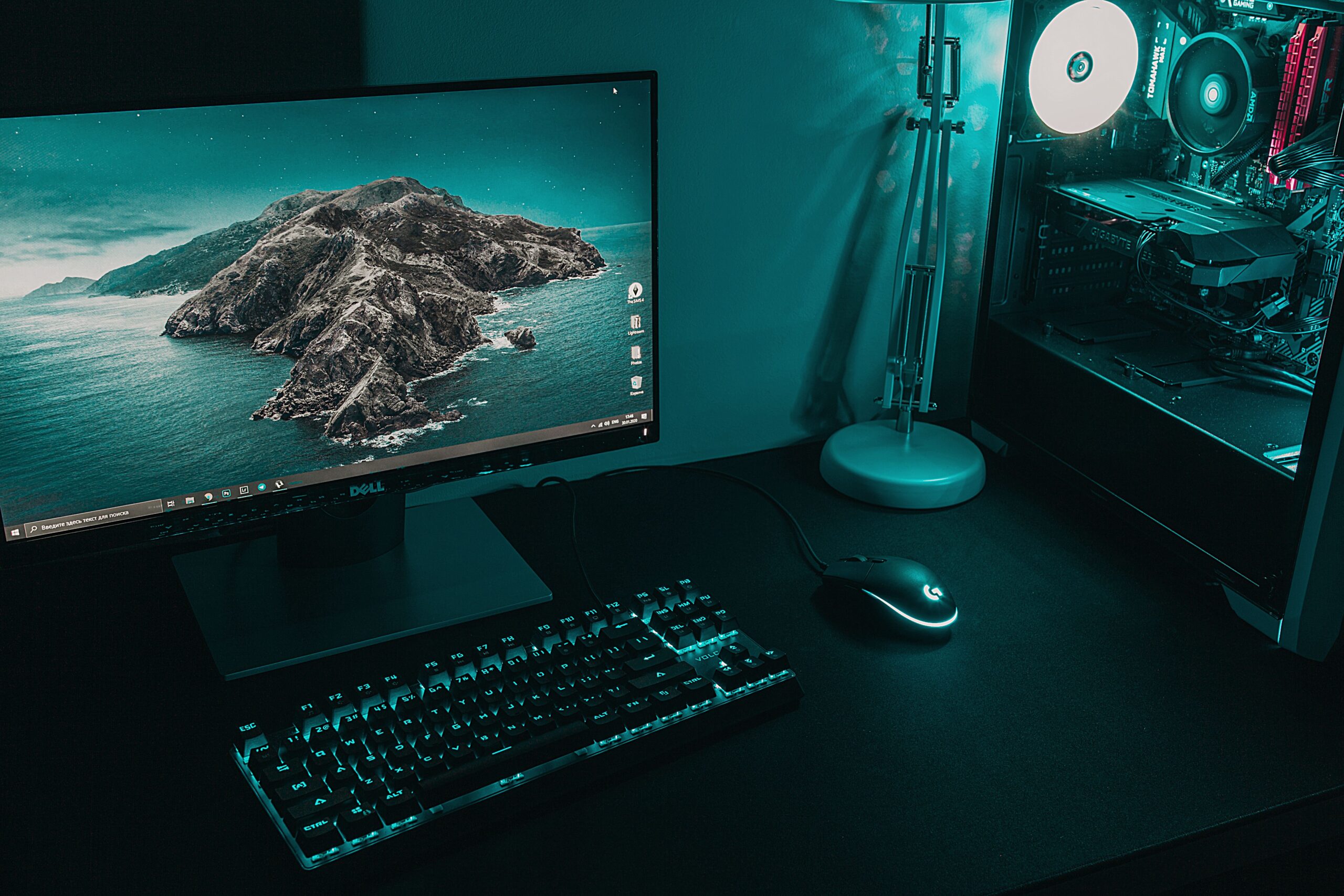 Desktop workstation with a wide monitor displaying a rocky island wallpaper a backlit teal keyboard and mouse and a teal lit PC tower on the right