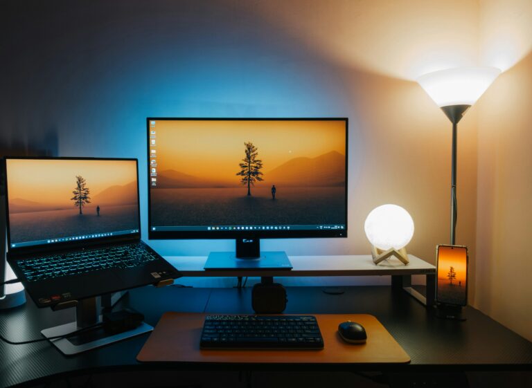 Desk setup with a laptop and a large external monitor showing a sunset scene; a floor lamp, smartphone on a stand, and keyboard/mouse on the desk.