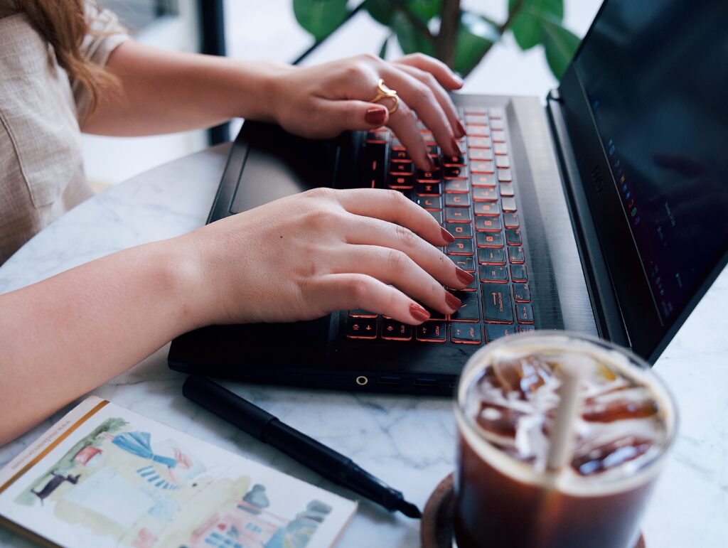 Close up of hands typing on a laptop with red backlit keys iced coffee in the foreground and a plant in the background