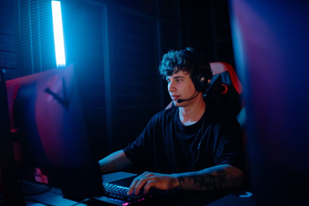 Young man wearing a headset and typing on a backlit keyboard in a dim room with blue neon lighting focused on the screen